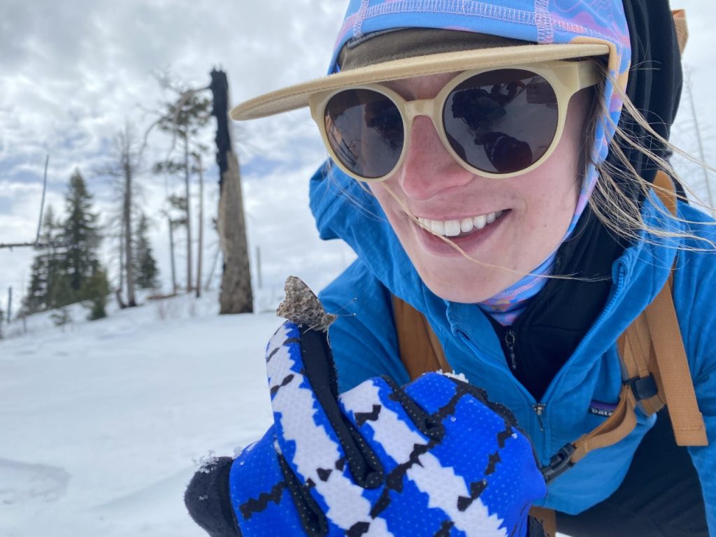 Female grad student in snow gear looks at moth perched on her glove. Background features snow, spruce trees, and partial clouds.